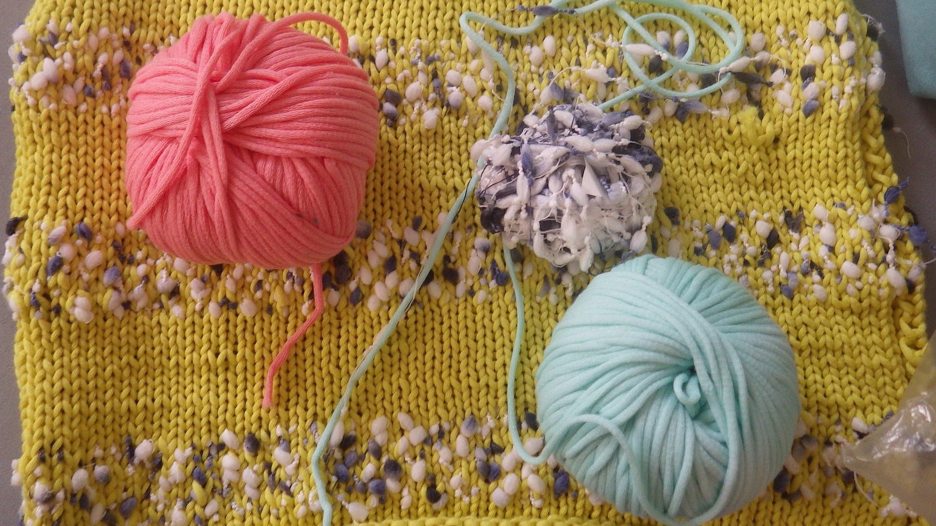 A piece of knitting is in the background. It's yellow with stripes of textured white and grey stripes. The ball of white and grey textured yarn sits on top of the knitting along side a ball of coral yarn and a ball of pale turquoise.